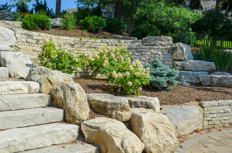 Limestone step and wall terrace with large boulders, mulch beds, evergreen shrubs, and perennial plantings.